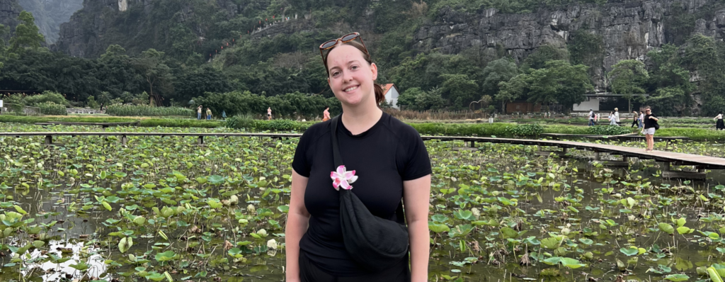 Smiling English teacher standing on a wooden walkway over a lotus pond with green mountains in the background, representing how teaching English abroad changes you through travel and new experiences.