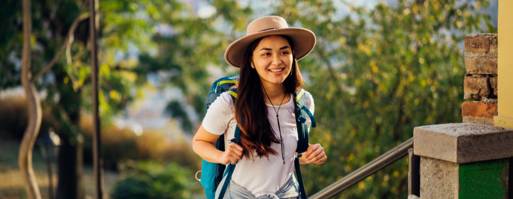 Smiling solo traveller with a backpack and hat walking through a sunny city, representing the safest countries to teach English abroad solo for TEFL teachers.