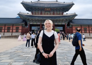 Happy TEFL teacher abroad standing in front of a grand Asian palace gate, enjoying cultural sightseeing on a day off from teaching in South Korea
